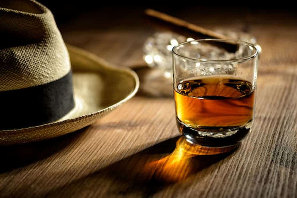 Half-full-glass-of-rum-on-wooden-table-with-cigar-and-hat.